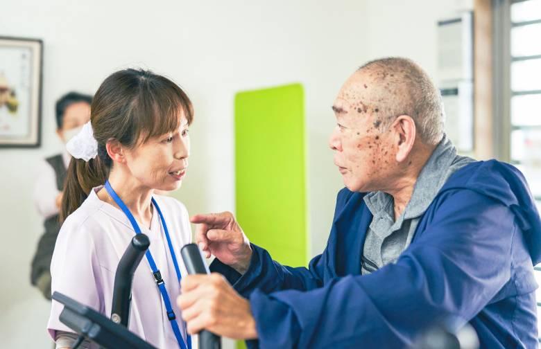 運動機器を使う利用者さんと目線を合わせて会話する、系列施設のスタッフ